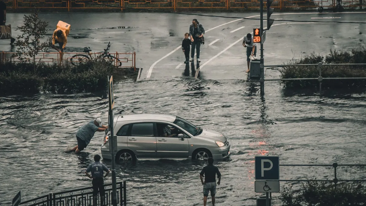 A car stranded in floodwater on a city street while people wade past